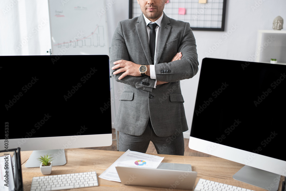 Naklejka premium cropped view of trader standing with crossed arms near laptop and computer monitors