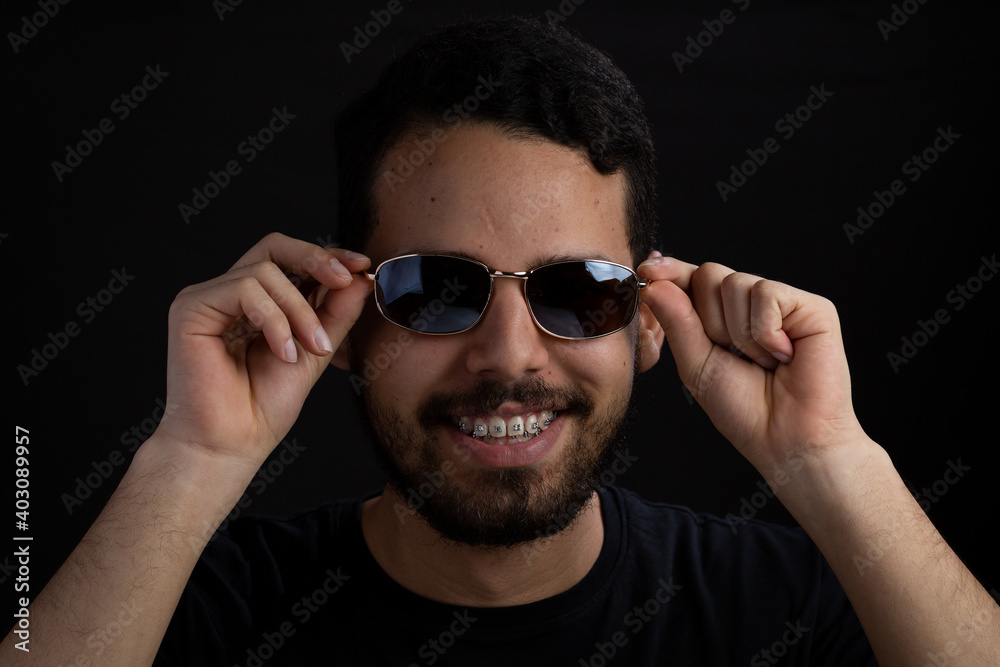 Obraz premium Young man frontal head shot with black background. He is smiling and taking off his sunglasses. He has a casual look with beard and t-shirt.