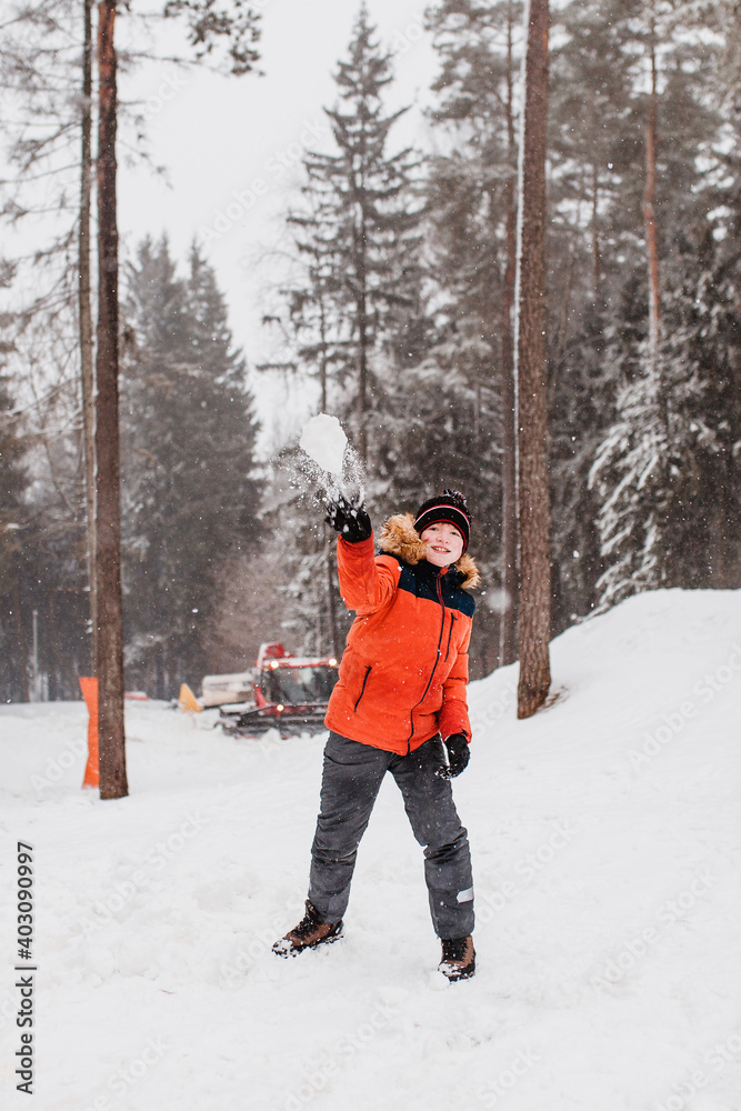 A boy in an orange winter jacket with a hood is having fun in the mountains throwing snowballs - New Year holidays