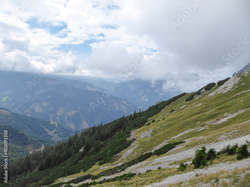 A panoramic view on the Austrian Alps from the Hochkogel peak. Thick clouds above the region. Endless mountain chains. The slopes are overgrown with small plants and trees. Mysterious landscape.