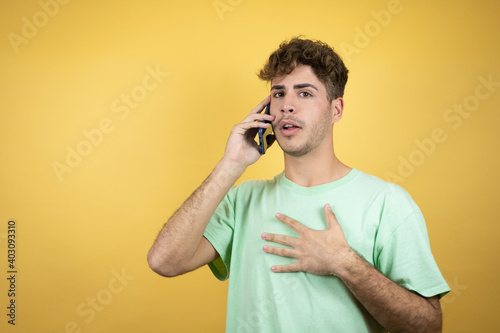 Handsome man wearing a green casual t-shirt over yellow background talking on the phone with a worried expression