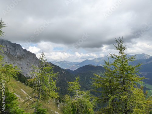 A panoramic view on the Austrian Alps from the Hochkogel peak. Thick clouds above the region. Endless mountain chains. Few pine trees in front, green slopes. Mysterious landscape. Beauty of the nature photo