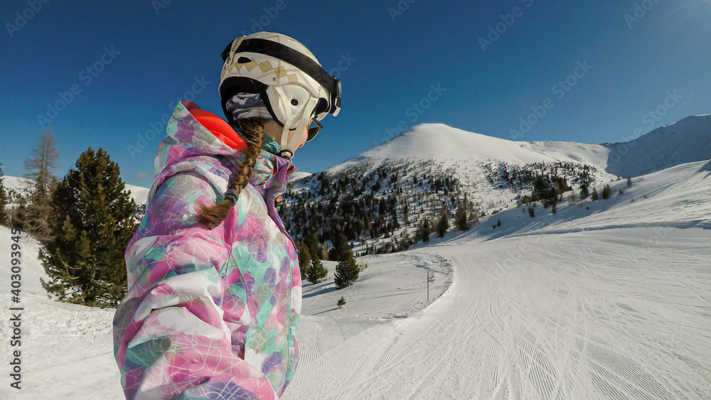A snowboarding woman in pink outfit taking a selfie while going down the slope in Innerkrems, Austria. Everything is covered with fresh snow. High mountains in the back. Winter outdoor activities