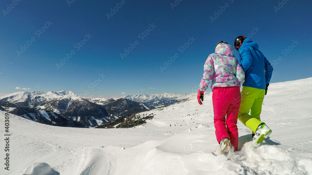 A couple in colorful skiing outfits and helmets walking on the fresh snow on top of Innerkrems, Austria. Everything is covered with fresh snow. High mountains in the back. Having fun and exploring A couple in colorful skiing outfits and helmets walking on the fresh snow on top of Innerkrems, Austria. Everything is covered with fresh snow. High mountains in the back. Having fun and exploring