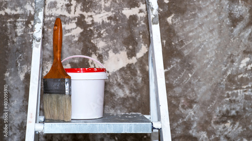 A paintbrush and a bucket of paint stand on a metal stepladder against an untreated concrete wall with remnants of white paint. Background photo