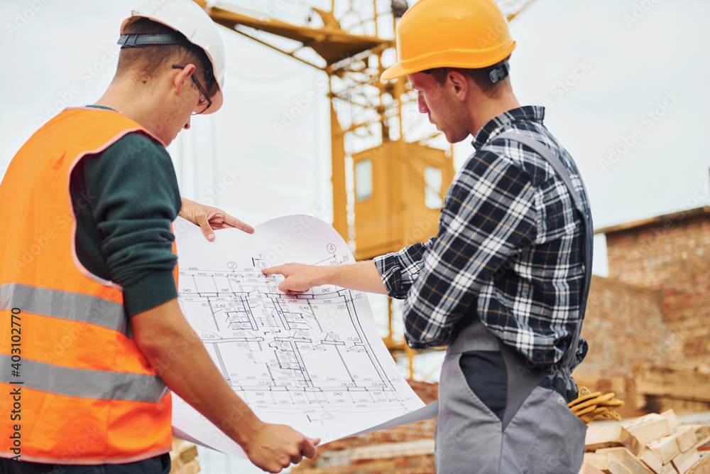 Using paper document with plan on it. Two construction workers in uniform and safety equipment have job on building together Using paper document with plan on it. Two construction workers in uniform and safety equipment have job on building together