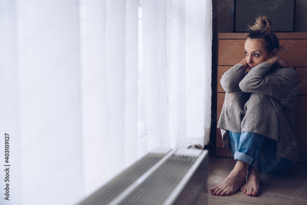 Young sad woman sitting on the floor in her living room looking out the window. Depression and despondency. Young sad woman sitting on the floor in her living room looking out the window. Depression and despondency.