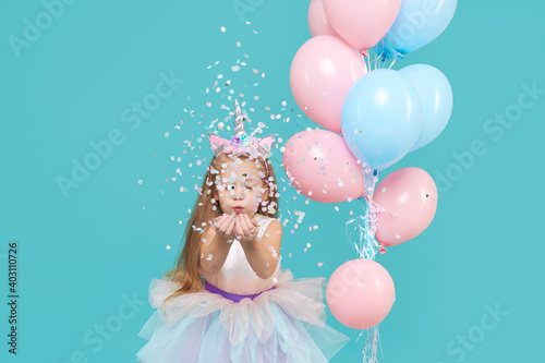 Obraz na płótnie Unicorn girl. Joyful child girl in elegant tulle dress and unicorn headband celebrates birthday party and blowing multicolored confetti on cyan background w salonie