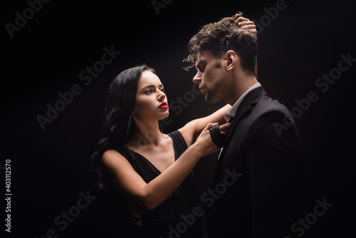 brunette woman with red lips looking at man and pulling hair isolated on black