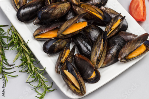 Mussels with open shells on white plate. Rosemary and slice of tomatoes on table