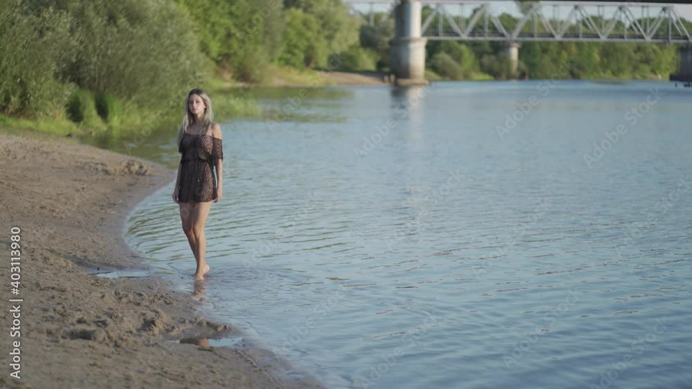 A cute blonde with long hair walks along the water's edge in a transparent tunic A cute blonde with long hair walks along the water's edge in a transparent tunic