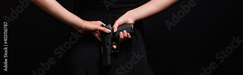 partial view of dangerous woman holding gun behind back isolated on black, banner
