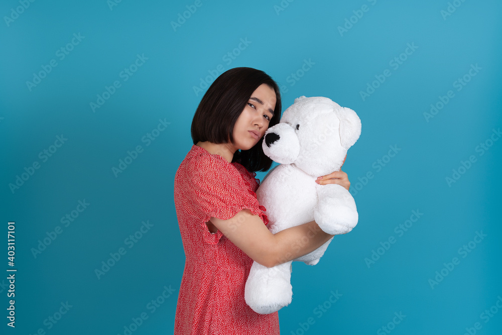 close-up portrait of a hurt, upset, pouting woman in a red dress clutching a white Teddy bear, isolated on a blue background. close-up portrait of a hurt, upset, pouting woman in a red dress clutching a white Teddy bear, isolated on a blue background.