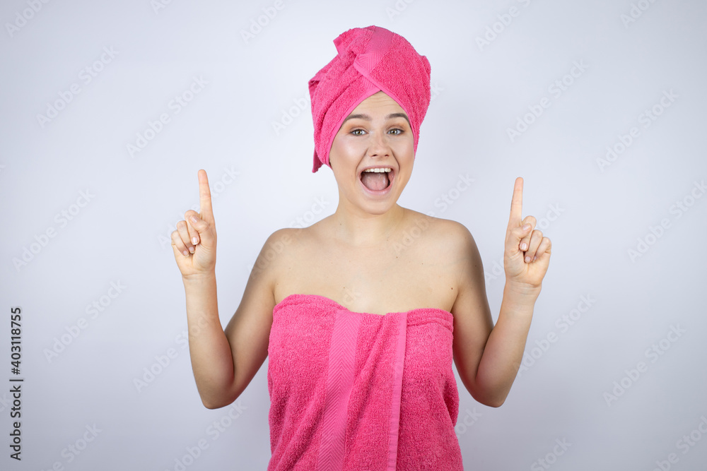 Young beautiful woman wearing shower towel after bath standing over isolated white background amazed and surprised looking at the camera and pointing up with fingers and raised arms Young beautiful woman wearing shower towel after bath standing over isolated white background amazed and surprised looking at the camera and pointing up with fingers and raised arms