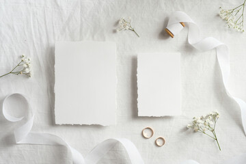 Blank wedding invitations cards mockups with space for text, golden rings, ribbon flowers on white textile background. Flat lay, top view.