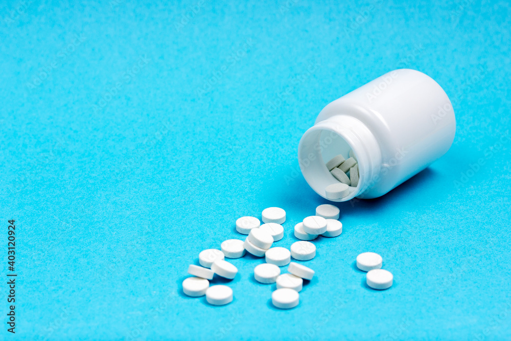 White, overturned medical jar with scattered pills on a blue background. Medications.