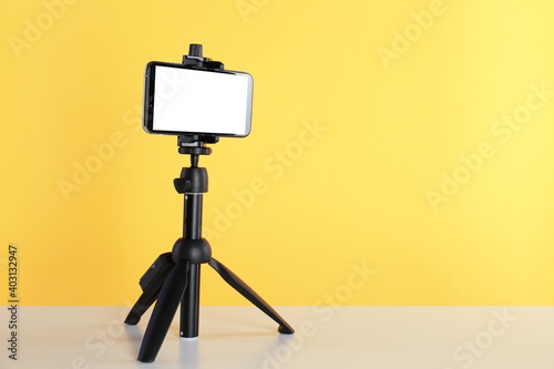 Smartphone with blank screen fixed to tripod on white table against yellow background. Space for text