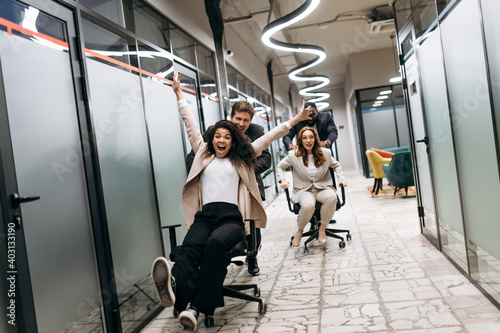 Young adult colleagues take a break from work. Friendly coworkers make a race on office chairs. Group of multiracial business people in formal wear having fun at the workplace