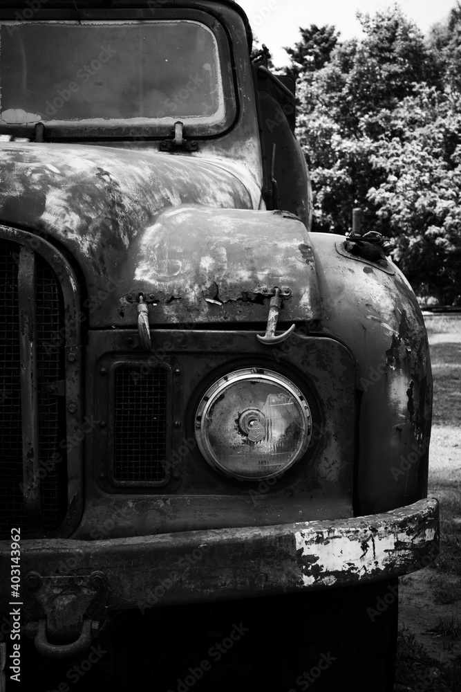Fototapeta premium Black and white detail of a vintage pick up truck showing head light and a section of grill. Truck has holes caused by rust. It has peeling, faded and scratched paint. The head light has broken glass