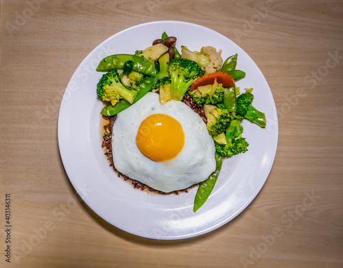 Cooked brown rice with fried vegetables and fried egg in white plate.