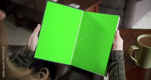 Over the shoulder slider shot of a young woman reading a book with green screen pages and a cup of coffee
