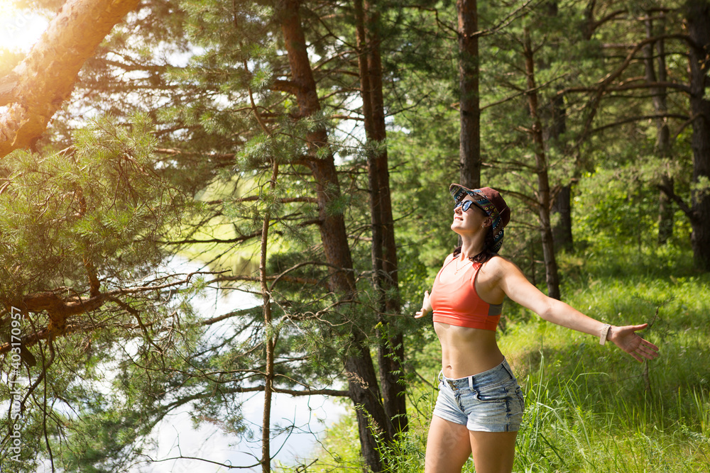 A woman in tourist sportswear, hat and sunglasses in the forest on the high bank of the river enjoys the sun, sunbathing, enjoying the beautiful view. Domestic tourism, ecotourism, unity with nature A woman in tourist sportswear, hat and sunglasses in the forest on the high bank of the river enjoys the sun, sunbathing, enjoying the beautiful view. Domestic tourism, ecotourism, unity with nature