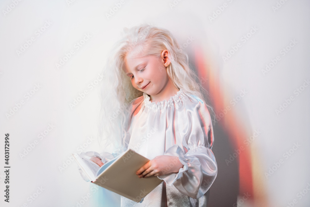 Child hobby. Kid leisure. Imagination development. Portrait of happy cute blonde albino little girl in white vintage dress reading favorite open book on light copy space background. Child hobby. Kid leisure. Imagination development. Portrait of happy cute blonde albino little girl in white vintage dress reading favorite open book on light copy space background.