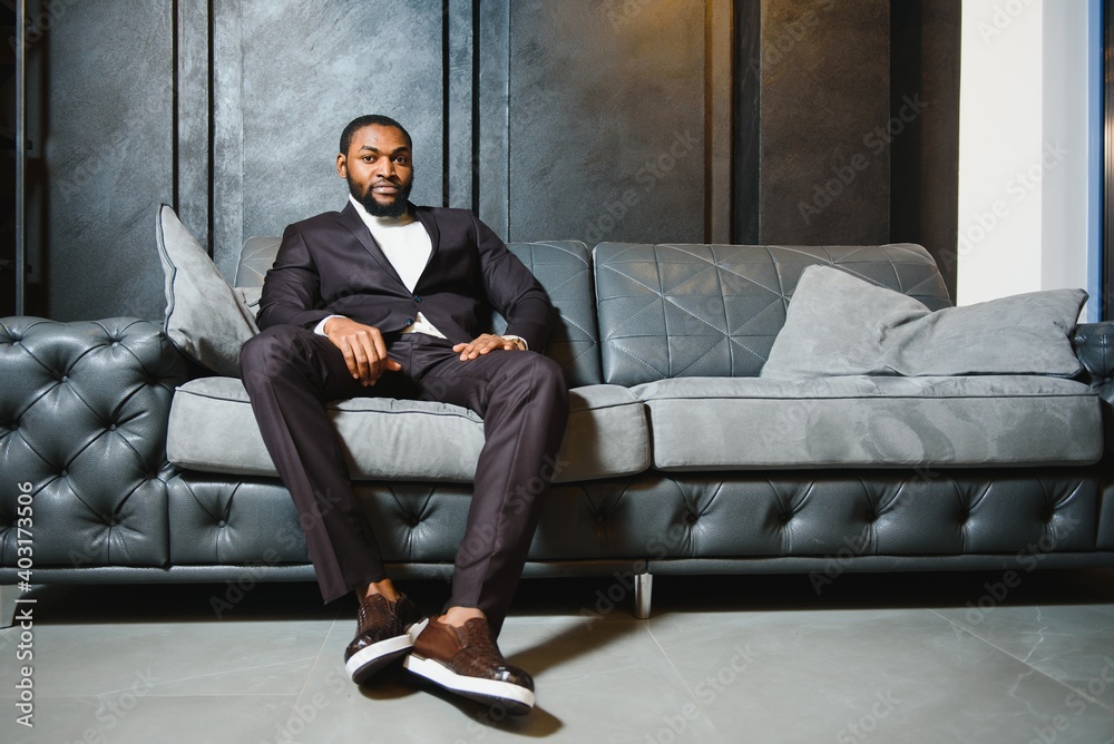 Young black man in suit on dark background sitting on a sofa. Fashion portrait of young man. Young black man in suit on dark background sitting on a sofa. Fashion portrait of young man.