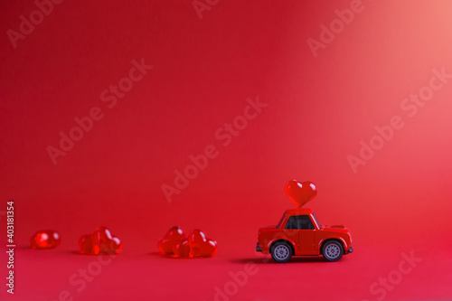Red toy car delivering a carriage of glass hearts on the roof against a red background. Valentine's day concept. Banner. Horizontal format. Copyspace