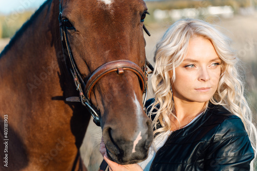 A pretty blonde in a black jacket stands in a field next to a horse at sunset.