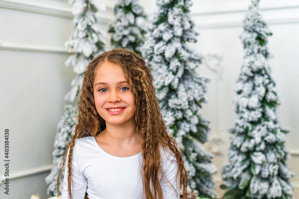 Happy smiling little girl sitting on the carpet by the Christmas tree at home waiting for gifts in the morning Happy smiling little girl sitting on the carpet by the Christmas tree at home waiting for gifts in the morning