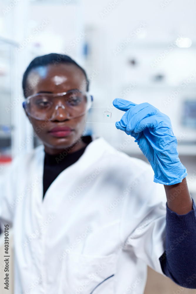 Close up of african biochemists holding microscope sample on glass slide with protective gloves. Black healthcare scientist in biochemistry laboratory wearing sterile equipment. Close up of african biochemists holding microscope sample on glass slide with protective gloves. Black healthcare scientist in biochemistry laboratory wearing sterile equipment.