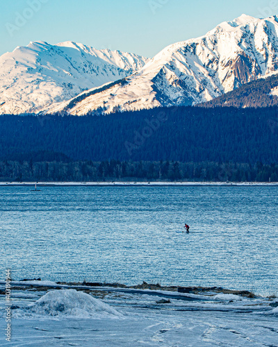 Standup paddle boarder in bay with snow and mountains.