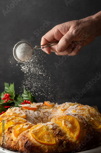 Close-up of woman's hand pouring sugar on homemade Roscon de Reyes, on dark background, in vertical