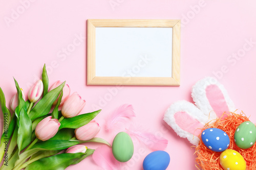 Easter eggs in a nest, bunny ears, tulips and a frame with copy space on a pink background. Flat lay greeting card.