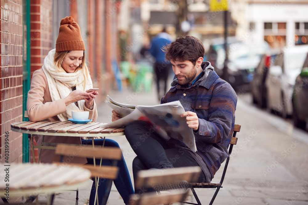 Couple On Date Sitting Outside Coffee Shop On High Street Using Mobile Phone And Reading Newspaper Couple On Date Sitting Outside Coffee Shop On High Street Using Mobile Phone And Reading Newspaper