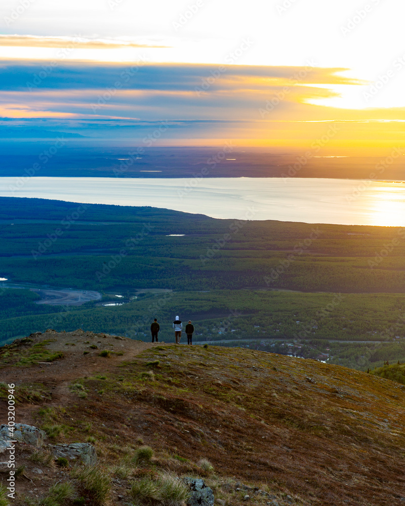 Three people stand atop a mountain overlooking a small town, an inlet, and a sunset.