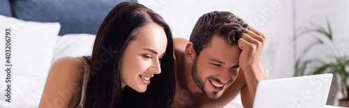 happy young couple laughing while looking at laptop on bed on blurred background, banner