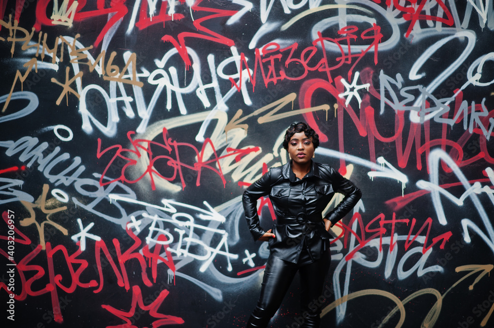 Fashionable beautiful african american woman posing in black leather jacket and pants at street against graffiti wall.