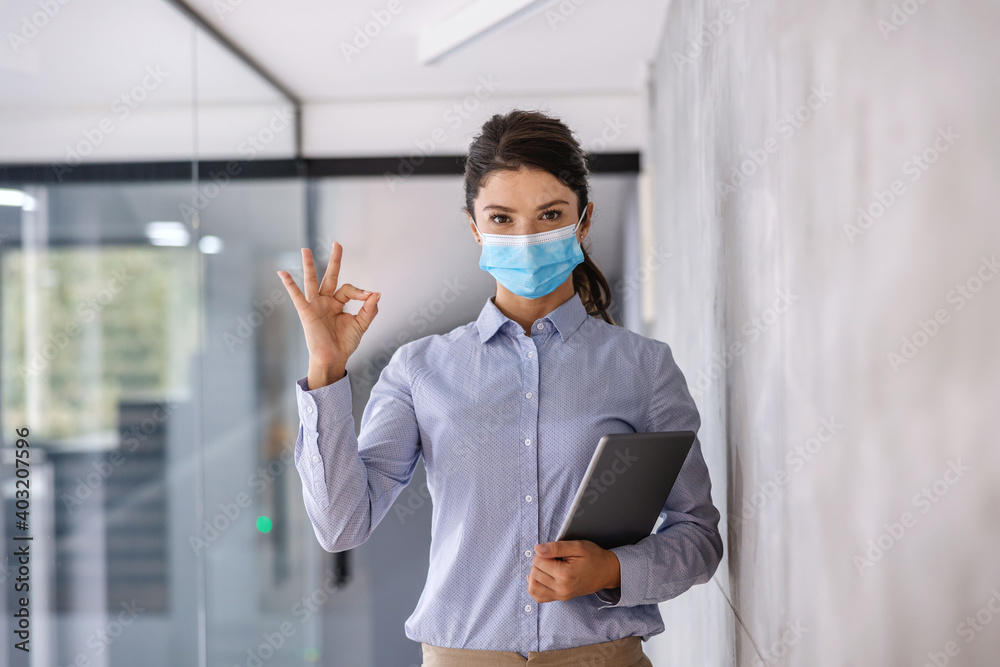 Young businesswoman with face mask standing in corporate firm with tablet in hands during corona virus and showing okay sign.
