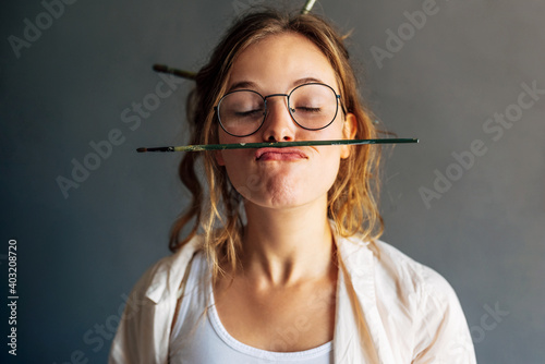 Closeup portrait of a funny beautiful female artist in eyeglasses holding with her lips a painting brush in her workshop. Pretty young woman painter making grimaces with a brush in her art studio.