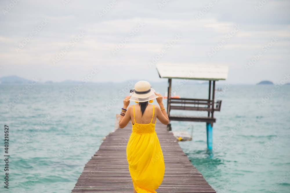 Close up portrait back young Asian woman in a yellow dress touching a hat on her head walking on a wooden bridge on the beach with a sky background