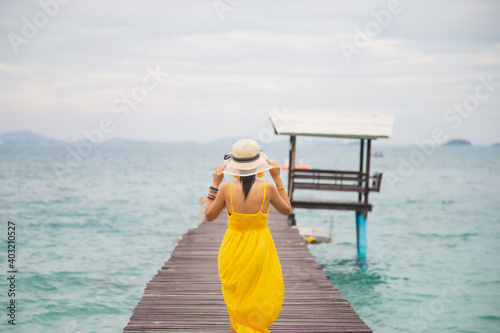 Close up portrait back young Asian woman in a yellow dress touching a hat on her head walking on a wooden bridge on the beach with a sky background © tisomboon