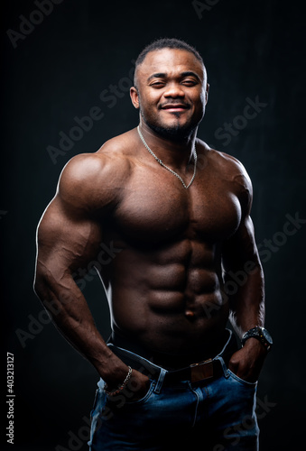 Young serious African American man with perfect muscular body posing to the camera in studio without shirt. Black background.