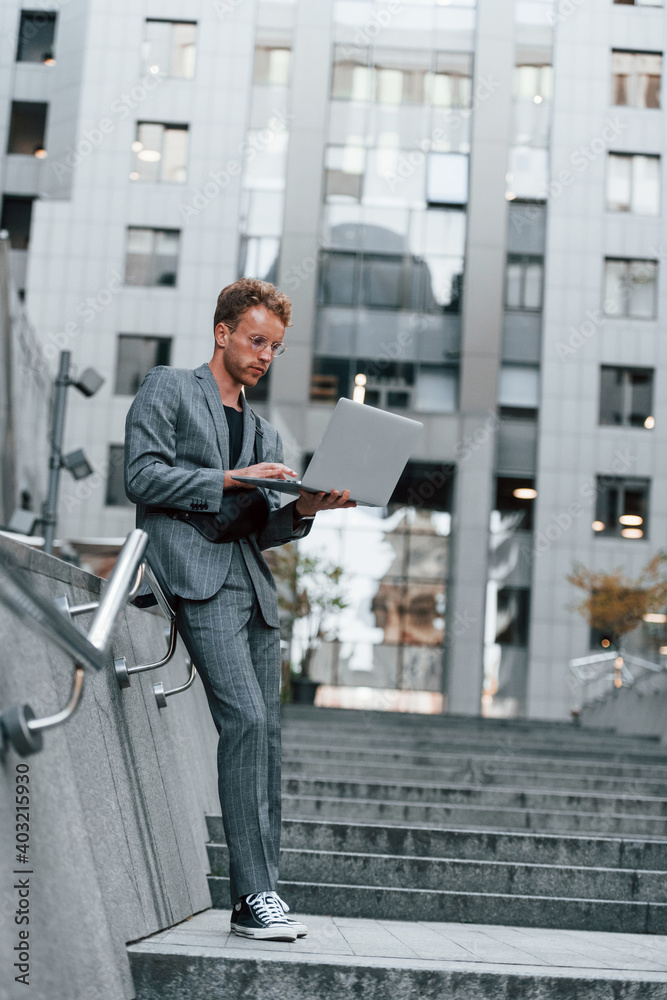 Laptop in hands. Young elegant man in good clothes is outdoors in the city at daytime Laptop in hands. Young elegant man in good clothes is outdoors in the city at daytime