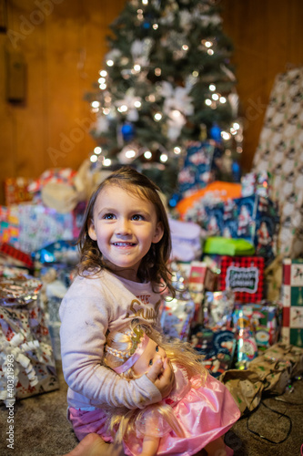 Happy girl holding a doll surrounded by presents and a Christmas tree