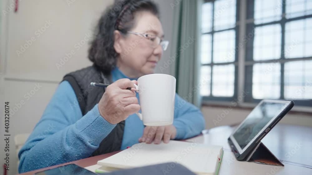 An old asian woman drinking hot drink from the mug while looking at online content on tablet screen, distanced learning, senior lady enjoy her afternoon adapting life to new digital modern technology An old asian woman drinking hot drink from the mug while looking at online content on tablet screen, distanced learning, senior lady enjoy her afternoon adapting life to new digital modern technology