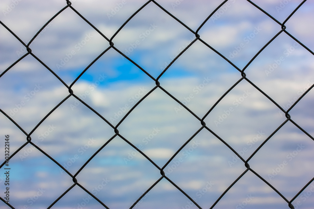 Metal protective grid or netting against a background of blue sky with white clouds. Protected facility, parking lot or prison. High quality photo Metal protective grid or netting against a background of blue sky with white clouds. Protected facility, parking lot or prison. High quality photo