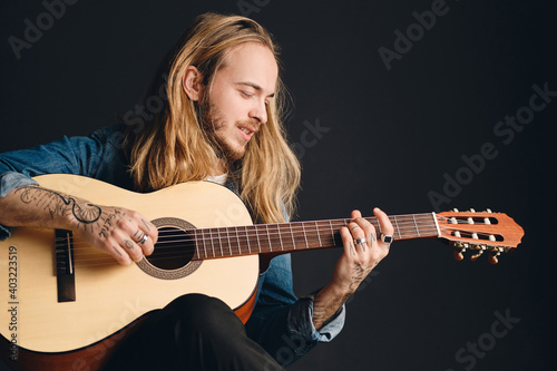 Portrait of long haired tattooed musician looking dreamy playing on acoustic guitar composing new song over black background
