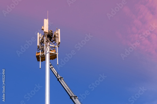 Fototapeta Naklejka Na Ścianę i Meble -  Workers install cellular base station with transmitters 3G, 4G, 5G and antennas on cell tower on background of pink-blue sky. Mobile telecommunication equipments. Copy space
